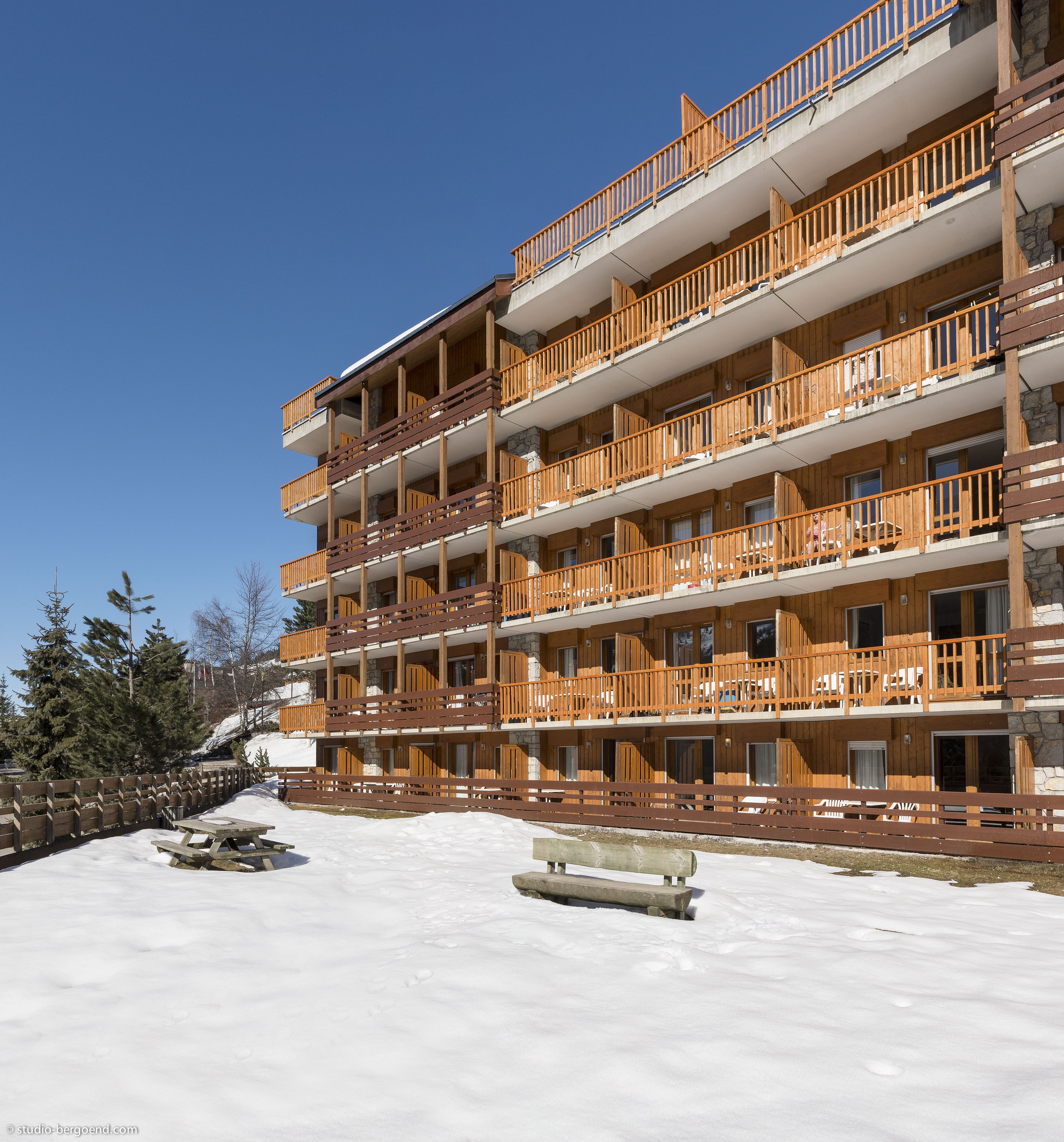 A multi-story wooden building with balconies, a snow-covered ground, and a clear blue sky.