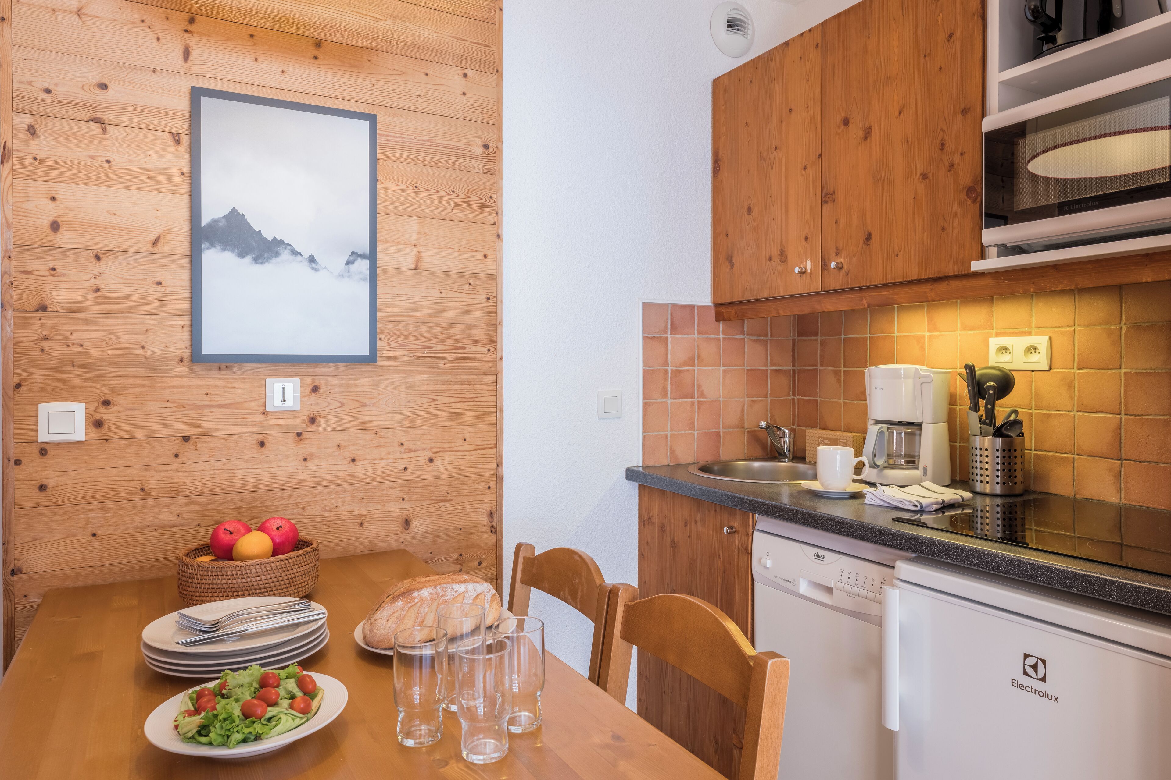 A wooden-paneled wall with a framed picture, a wooden dining table with plates and glasses, a basket of fruit, and a coffee maker on the counter.