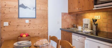 A wooden-paneled wall with a framed picture, a wooden dining table with plates and glasses, a basket of fruit, and a coffee maker on the counter.