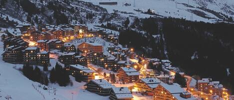 A snow-covered mountain town with multiple buildings, including lodges and hotels, and a ski slope in the foreground.