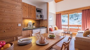 A cozy kitchen with wooden cabinets, a dining table set with plates, glasses, and a basket of fruit. A microwave is above the counter, and a window offers a view of a snowy landscape.