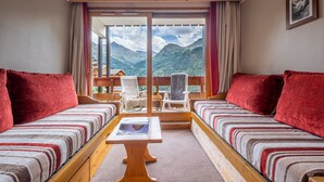 A room with two beds, a wooden table, and a view of mountains through large windows.
