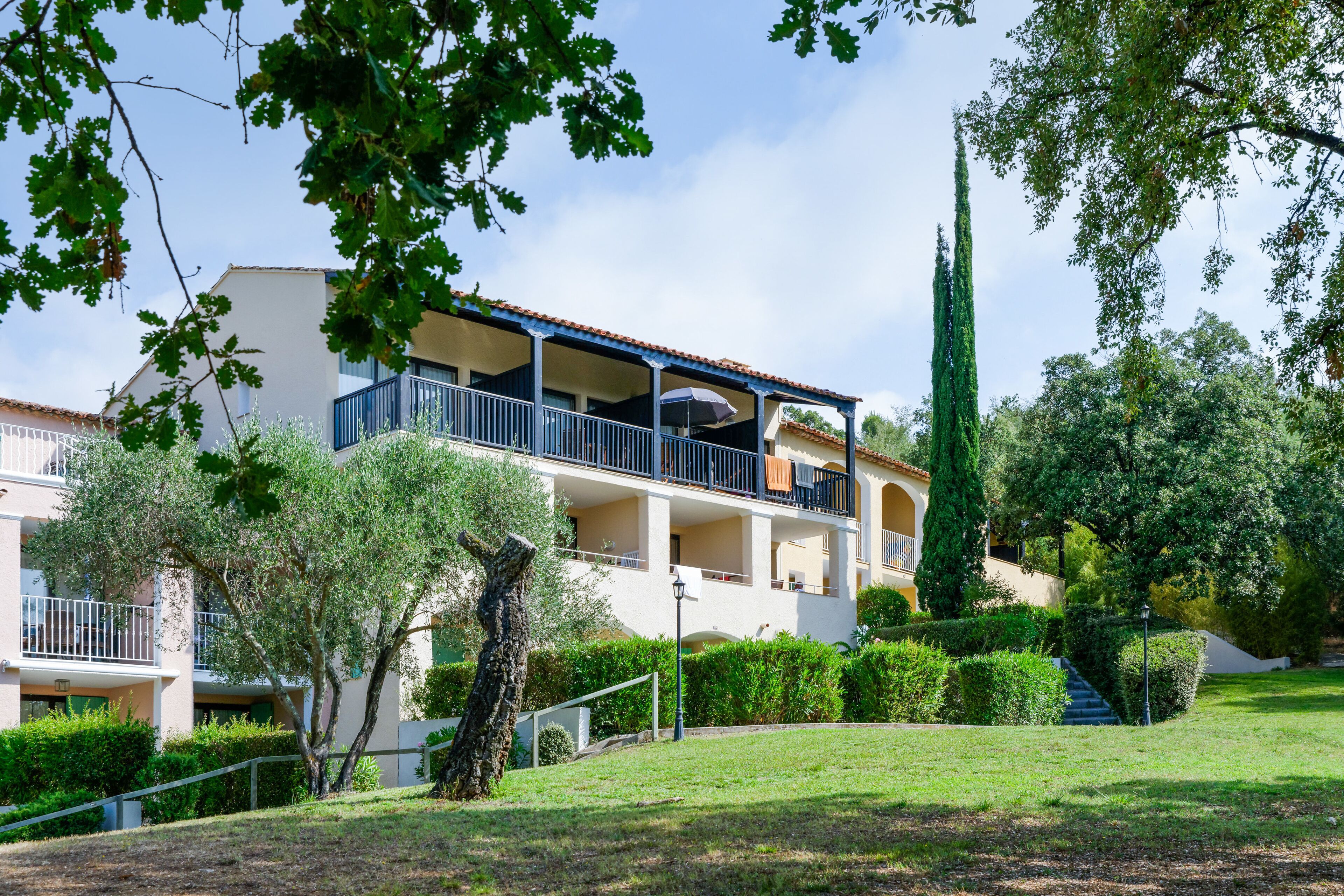 A multi-story building with balconies, surrounded by well-maintained greenery and a clear sky.