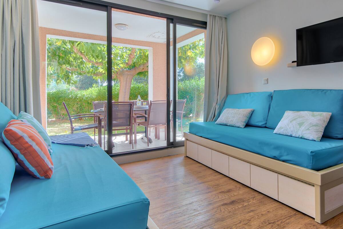 A modern living room with a sofa and a table, large sliding glass doors leading to an outdoor area with a tree and patio furniture.