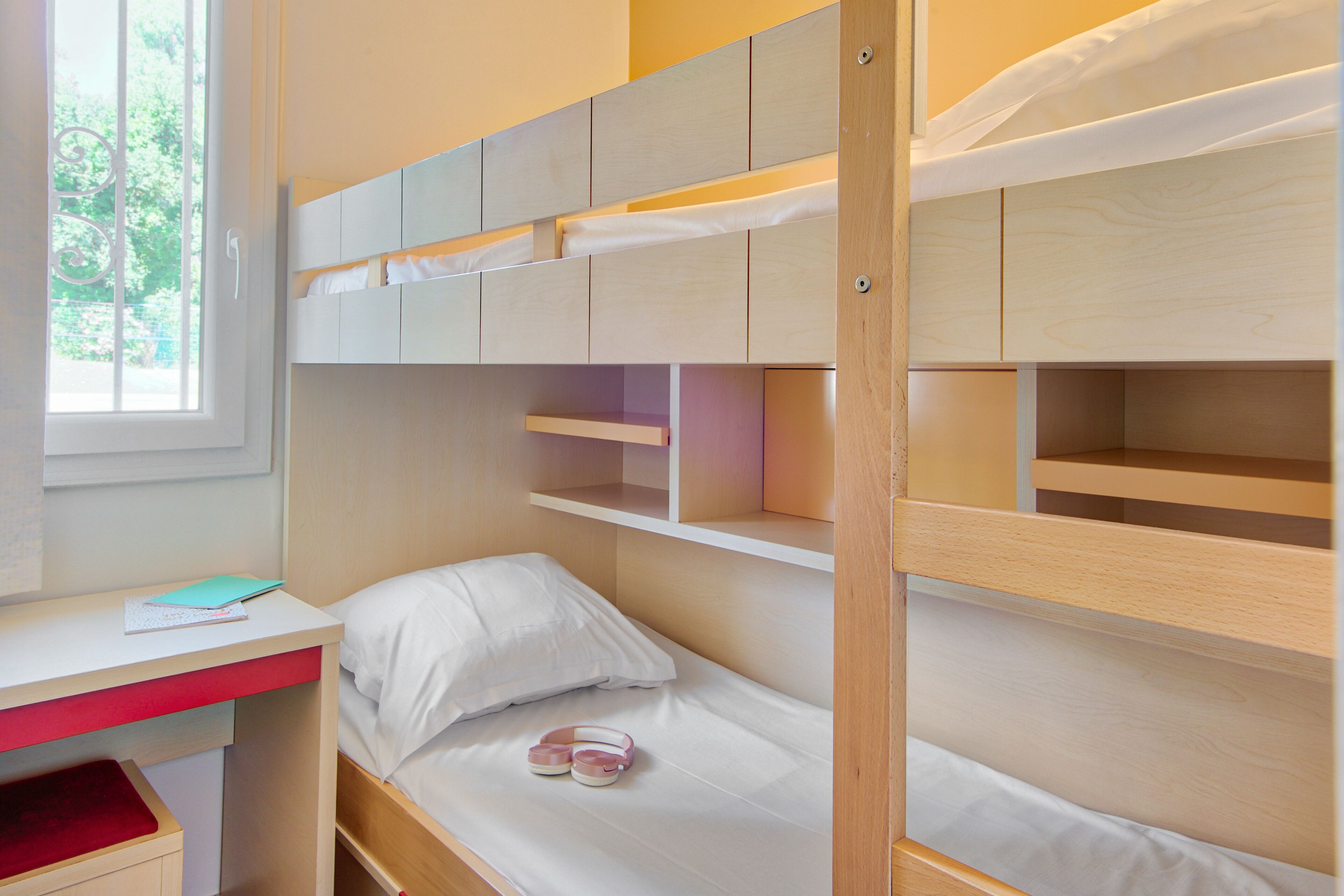 Una cama individual con escritorio y estanterías, una ventana con vistas a zonas verdes, y un par de zapatos rosas en la cama inferior.