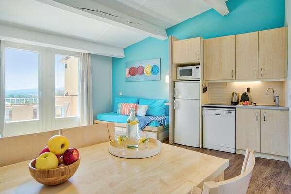 A modern kitchen with a microwave, dishwasher, and sink. A dining table with a wooden bowl of fruit is in the foreground. - Pierre & Vacances Résidence Les Parcs de Grimaud (Grimaud)