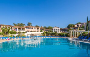 A large swimming pool with blue lounge chairs and a building with multiple balconies in the background. - Pierre & Vacances Résidence Les Parcs de Grimaud (Grimaud)