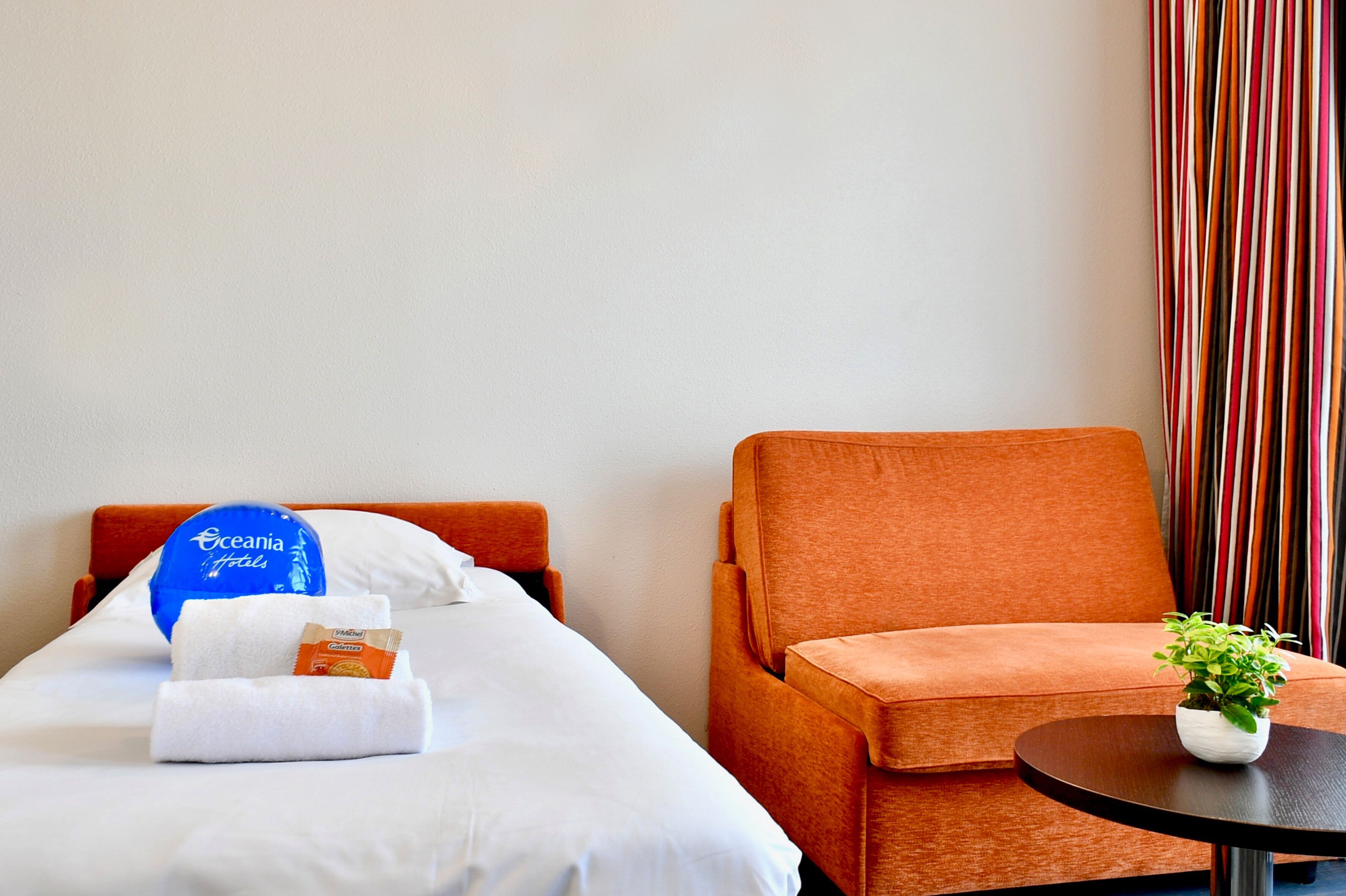 Una habitación de hotel con una cama, una silla, una mesita con una planta y una cortina a rayas.