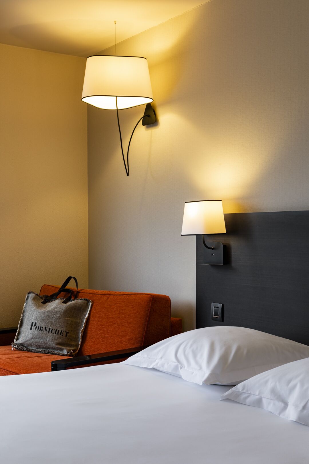 A hotel room with a bed, two wall-mounted lamps, an orange armchair, and a pillow with a bag labeled "Pornichet."