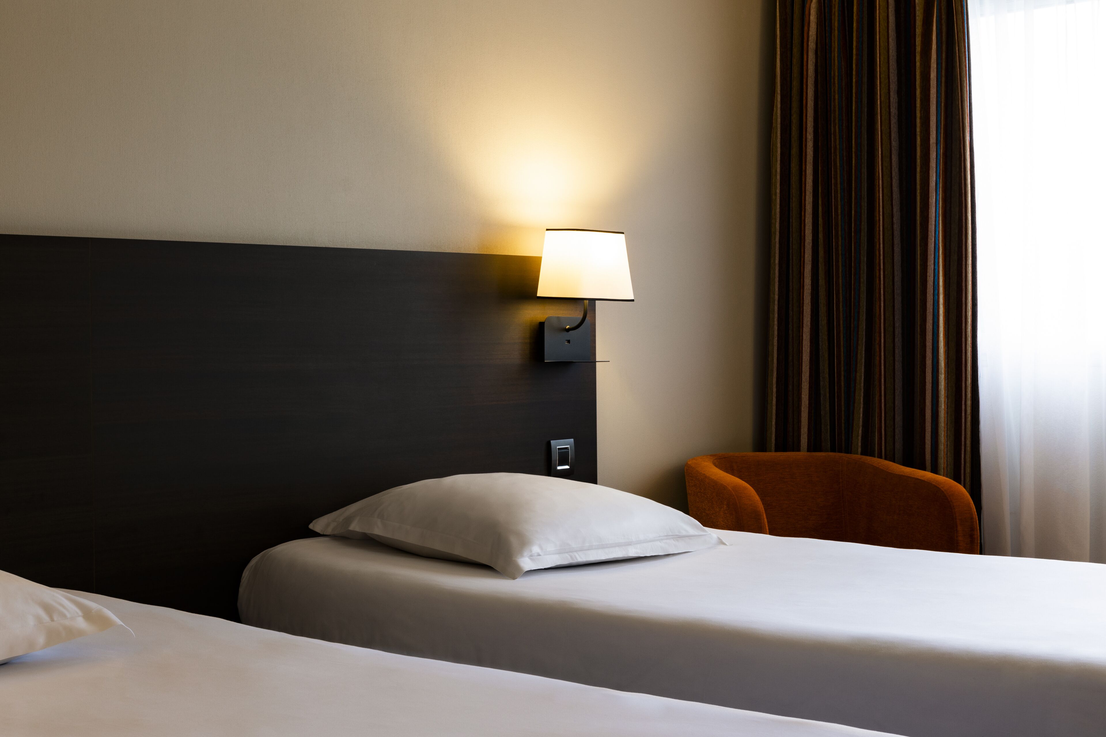 Une chambre d’hôtel avec deux lits, une lampe fixée au mur, une chaise et un rideau.
