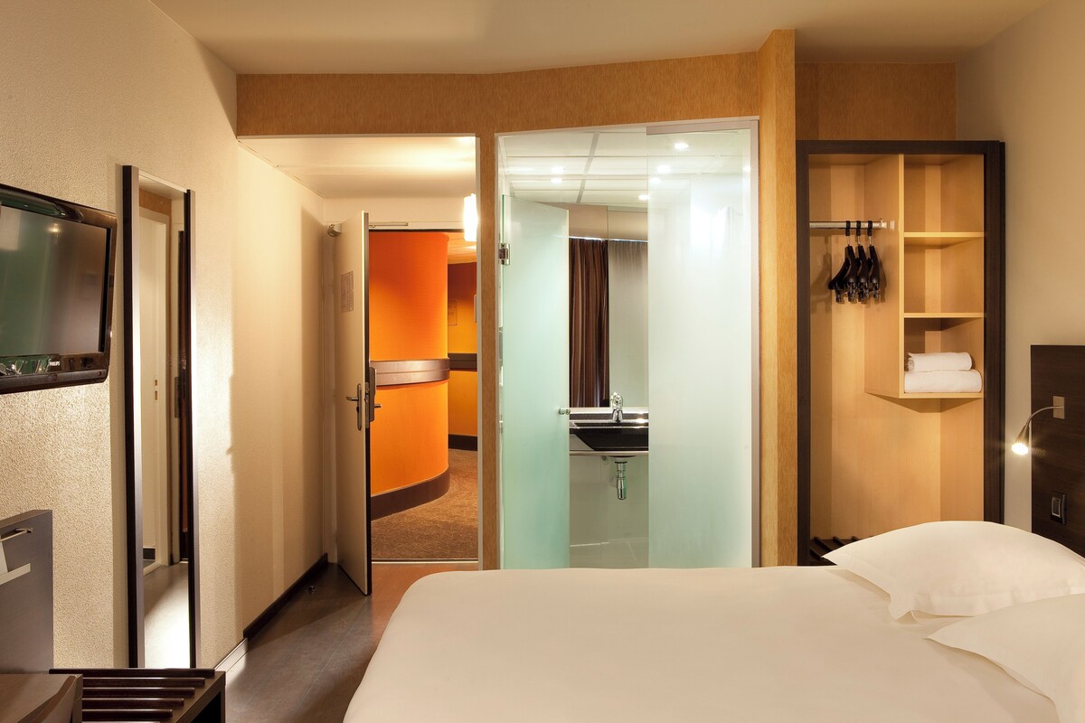 Une chambre d’hôtel avec un lit, une salle de bain avec un lavabo et une armoire avec des vêtements suspendus.