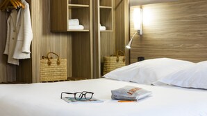 Une chambre d’hôtel avec une armoire en bois, un lit avec du linge de lit blanc, une lampe de chevet et un magazine sur le lit.