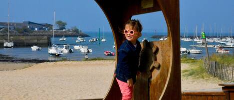 A child standing in a wooden structure at a beach with boats in the background.