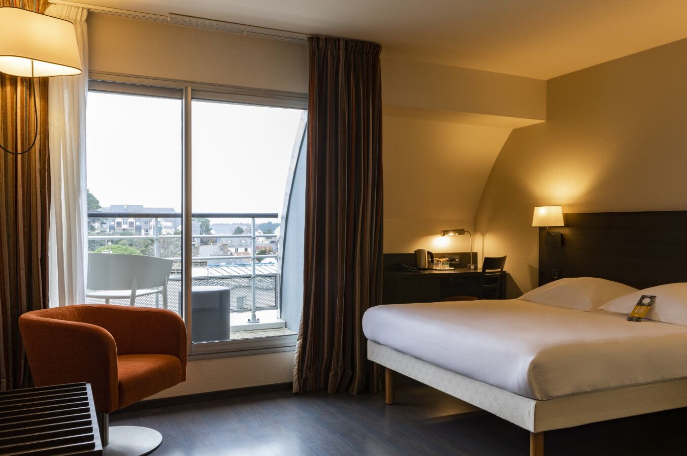 A hotel room with a bed, a chair, a desk with a lamp, a window with curtains, and a view of the city.