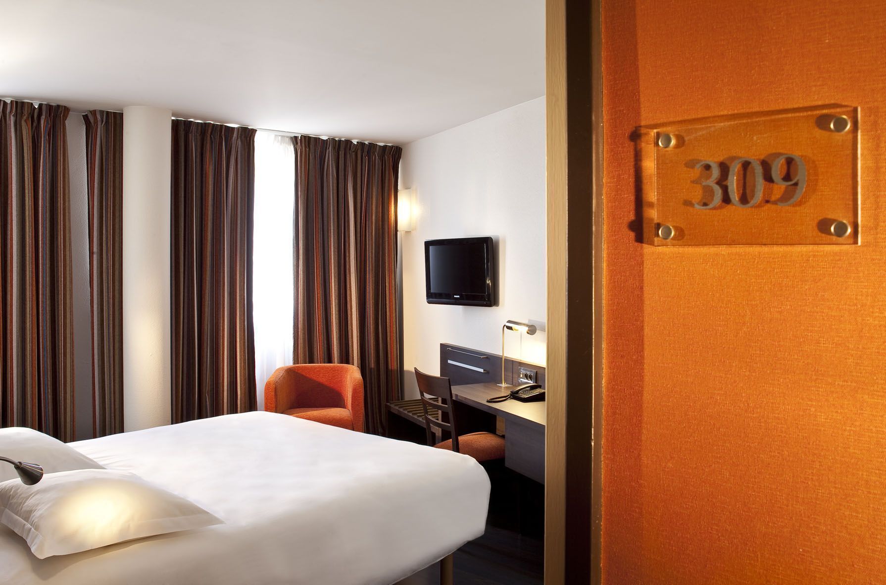 A hotel room with a bed, a desk with a chair, a TV, and curtains.