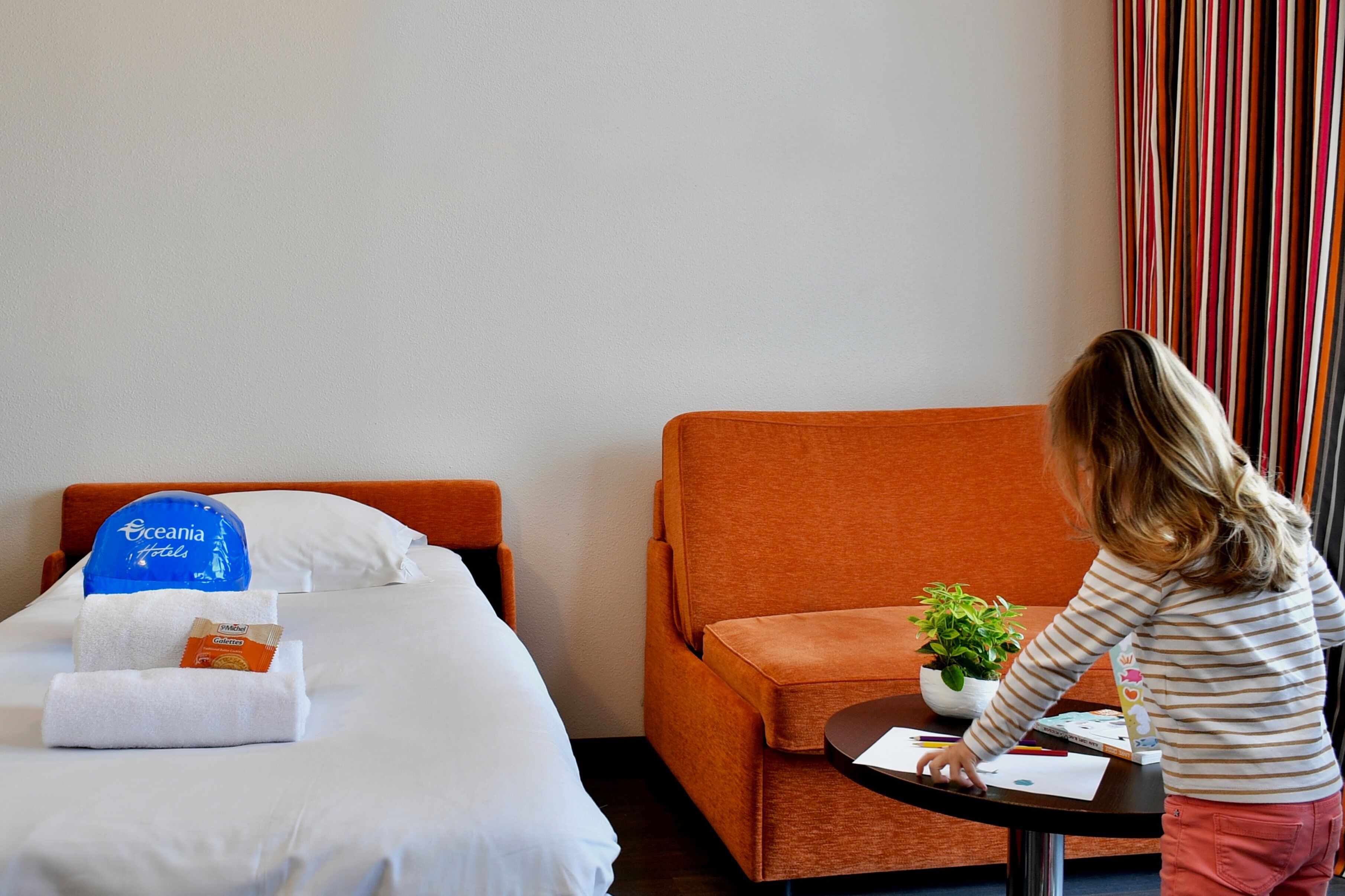 A hotel room with a bed, a small orange armchair, a table with a plant, and a child drawing.