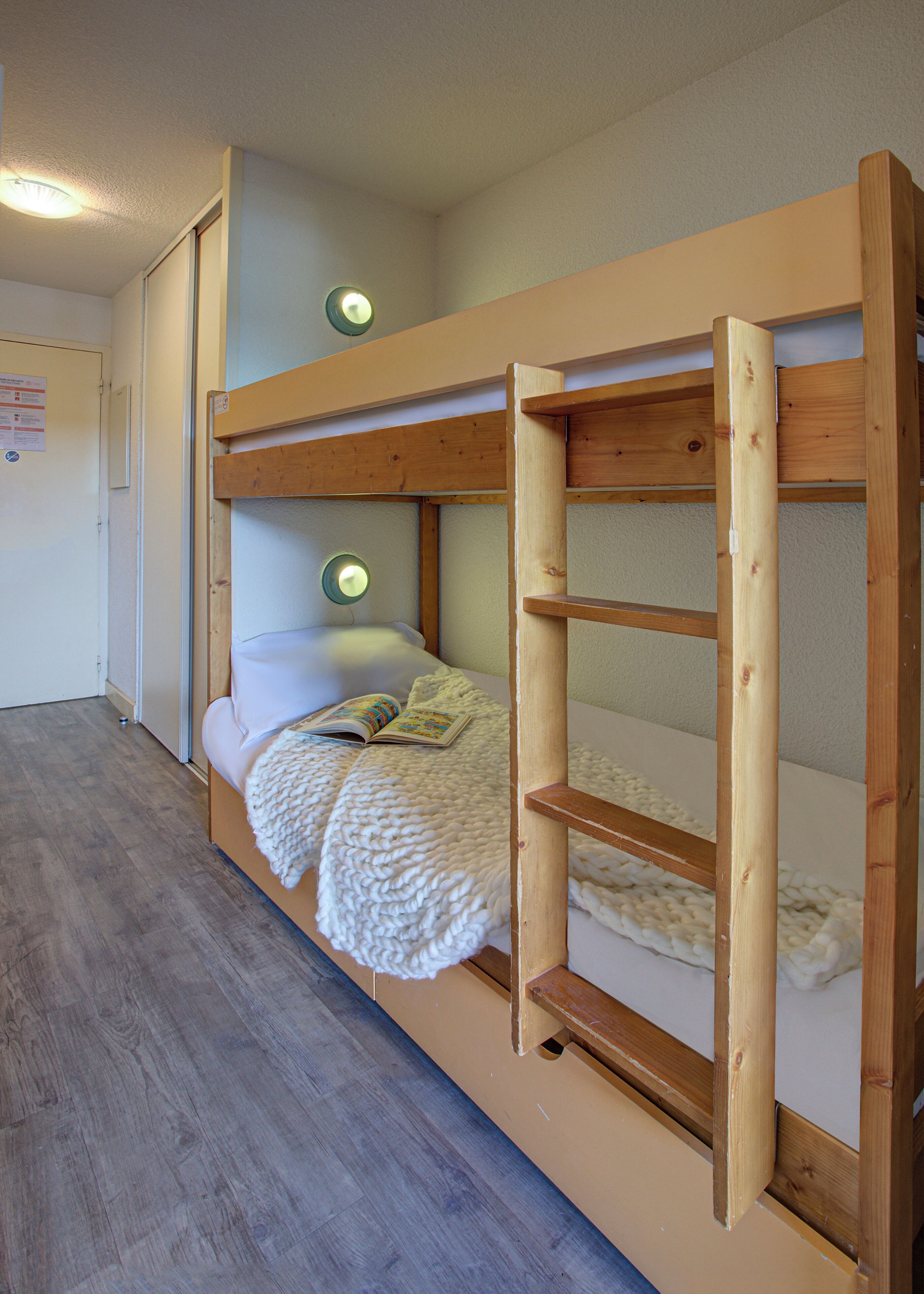 Una cama litera con estructura de madera, un colchón blanco y una manta blanca. Hay una mesita con una lámpara y un libro encima.