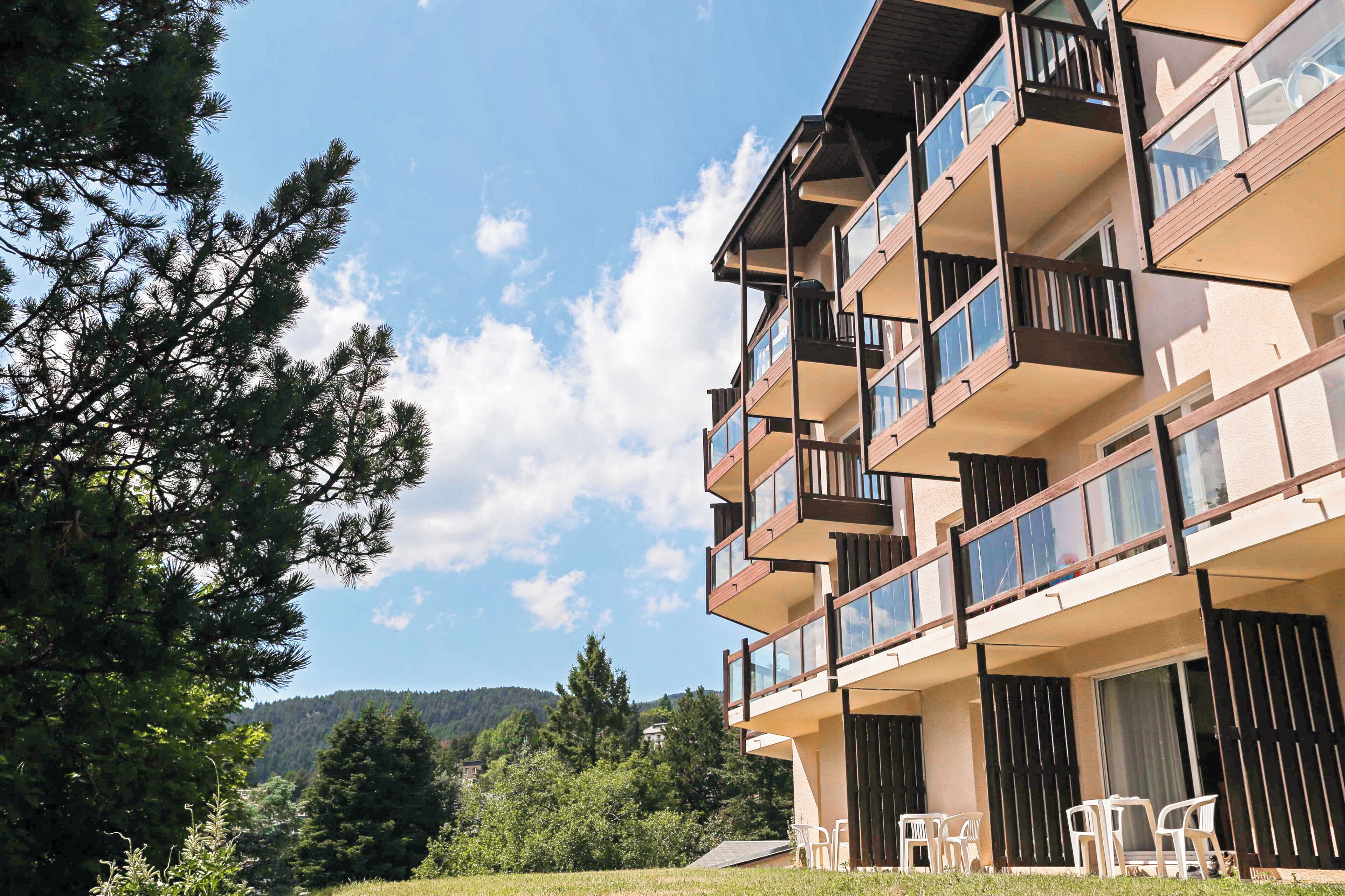 Un edificio de varios pisos con balcones, mobiliario exterior y vistas a árboles y colinas.
