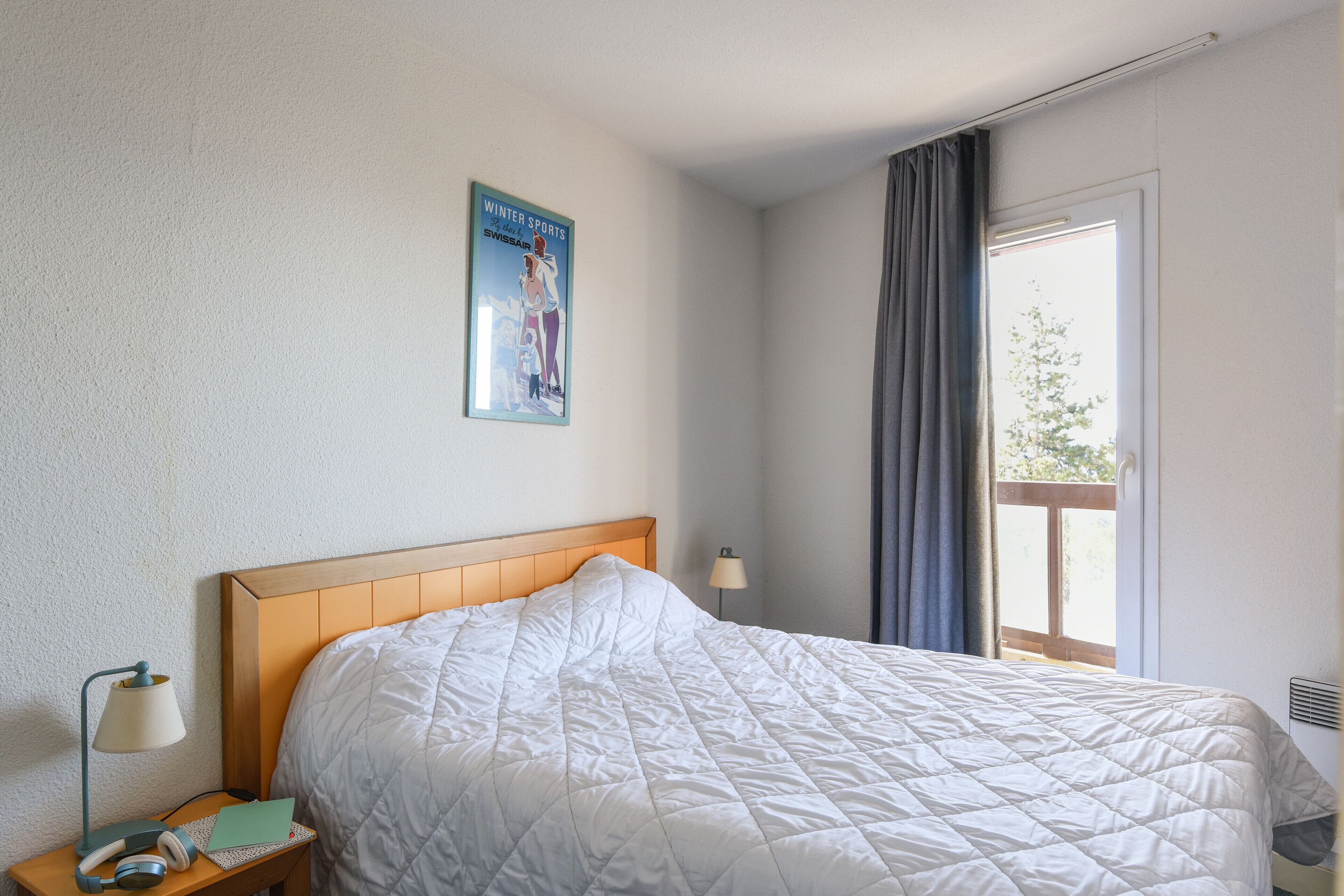 A hotel room with a bed, a nightstand, a lamp, a poster on the wall, and a window with curtains.