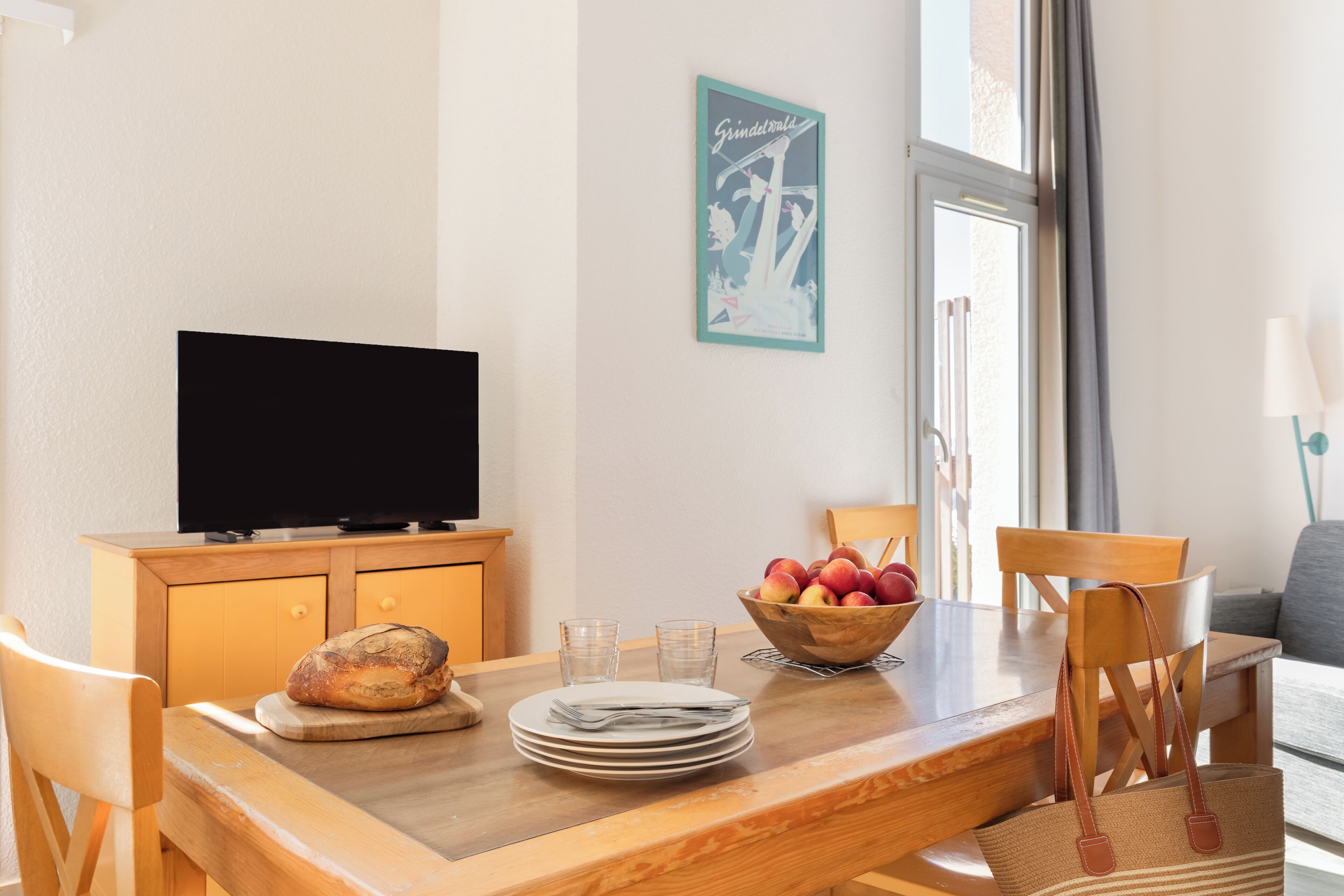 A dining area with a wooden table, chairs, a television, a bowl of fruit, and a poster on the wall.