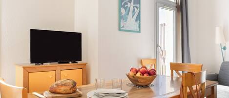A dining area with a wooden table, chairs, a television, a bowl of fruit, and a poster on the wall.