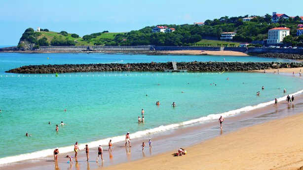 A beach with people swimming and walking along the shore, a rocky breakwater, and houses on a hill in the background. - Pierre & Vacances Résidence Eguzki (Saint-Jean-de-Luz)