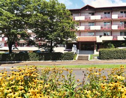 Un hotel de varios pisos con balcones, tejado de tejas rojas y un jardín bien cuidado con flores amarillas.