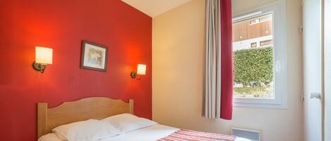 Habitación de hotel con cama, colcha a rayas rojas, dos lámparas de lectura fijadas a la pared y una ventana con cortinas.