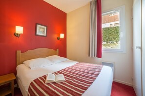 Quarto de hotel com uma cama, um cobreleito listrado de vermelho, duas luminárias de leitura fixadas na parede e uma janela com cortinas.