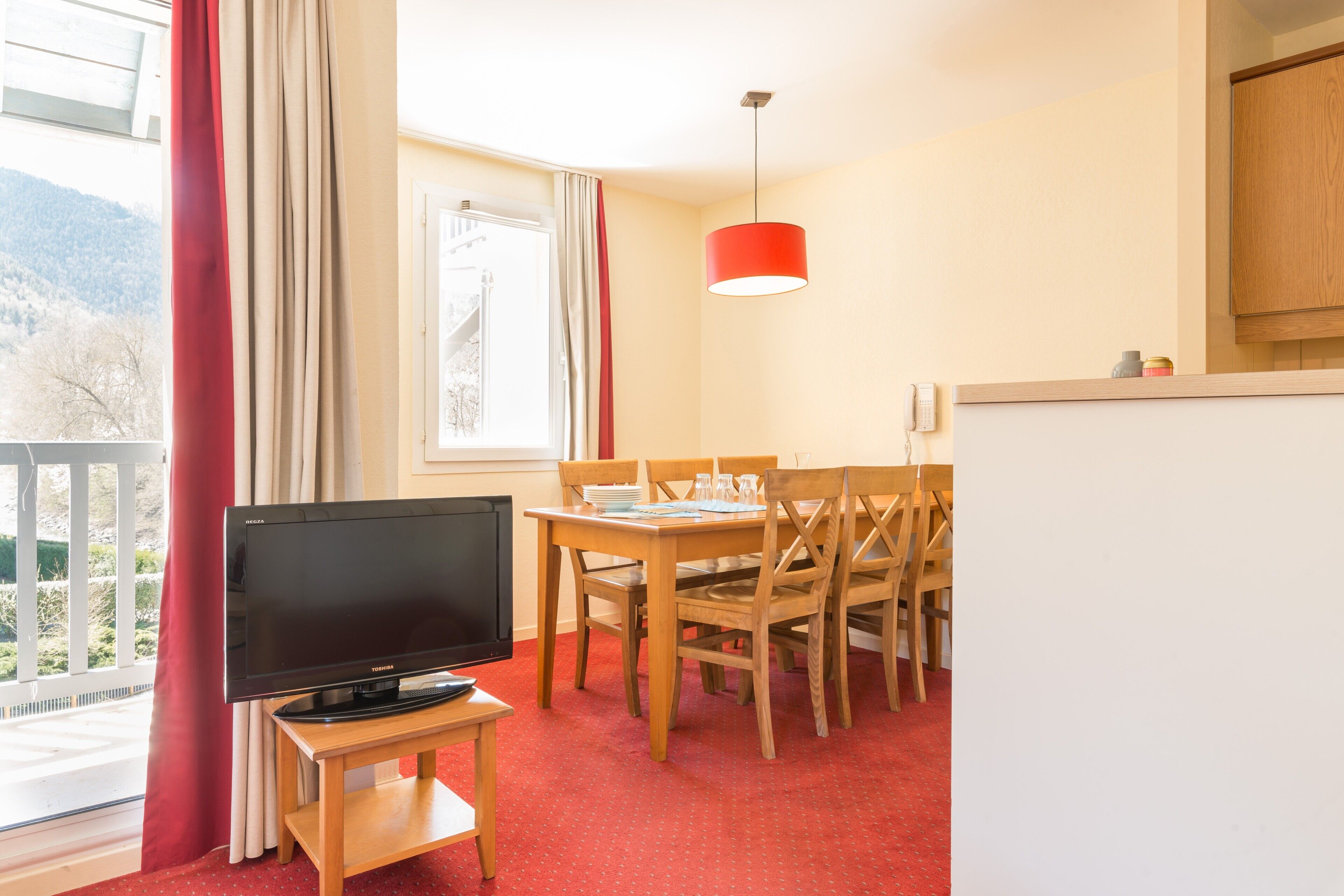 Una habitación con televisor sobre un mueble bajo, una mesa de comedor con sillas, una alfombra roja, un ventanal con cortinas y una lámpara colgante roja.
