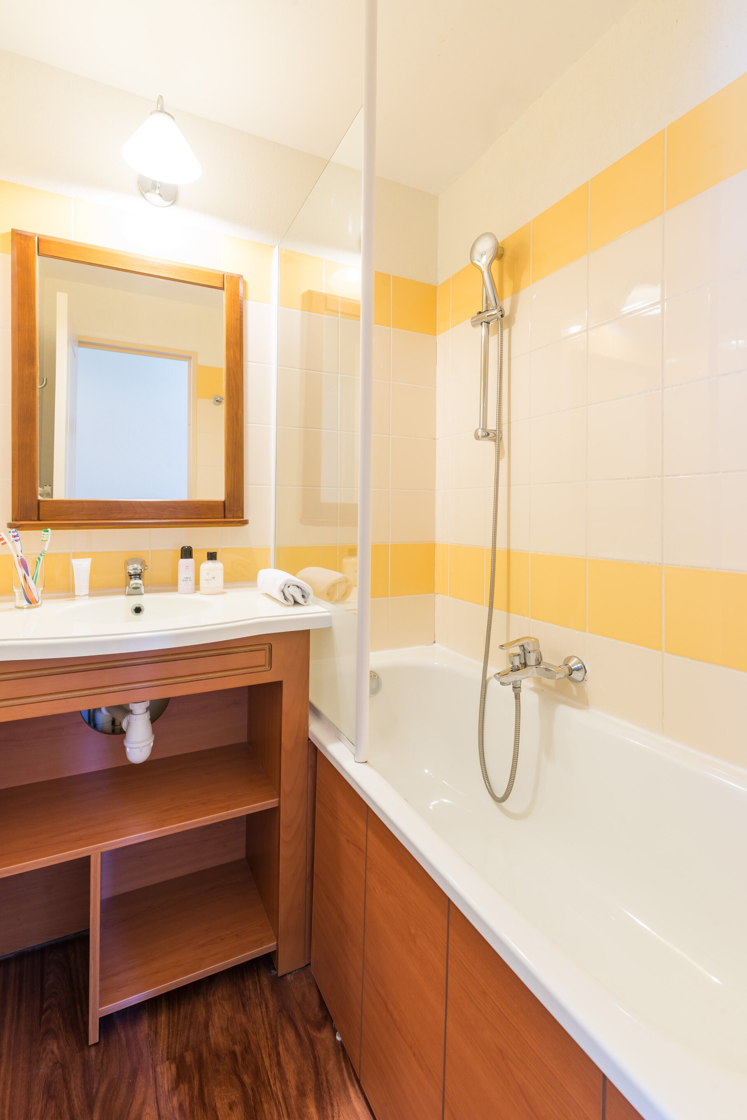 Un baño con tocador de madera, un lavamanos blanco, un espejo, una ducha con cabezal de ducha manual y paredes cubiertas de azulejos amarillos y blancos.