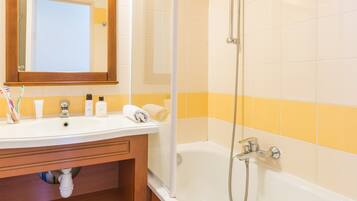 Un baño con tocador de madera, un lavamanos blanco, un espejo, una ducha con cabezal de ducha manual y paredes cubiertas de azulejos amarillos y blancos.