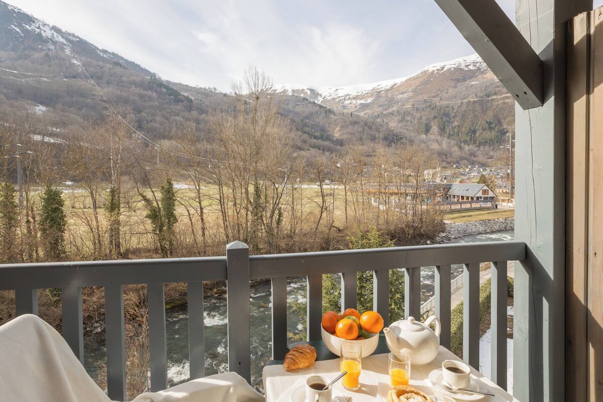 A breakfast table with a bowl of fruit, croissant, coffee, and juice, overlooking a river and mountains.