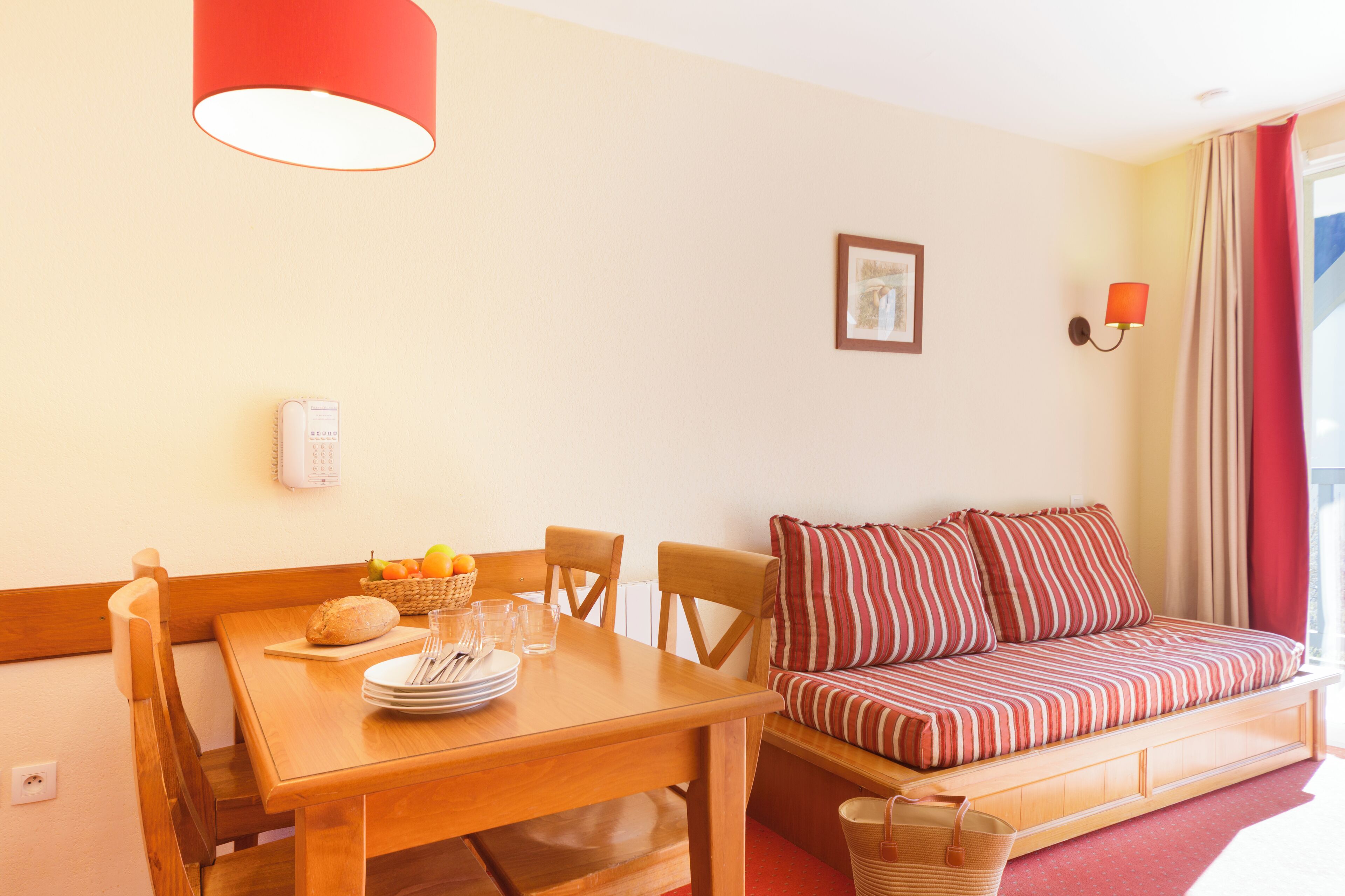 Een gezellige woonkamer met een houten eettafel gedekt voor twee personen, een gestreepte bank en een rode lamp aan de muur.