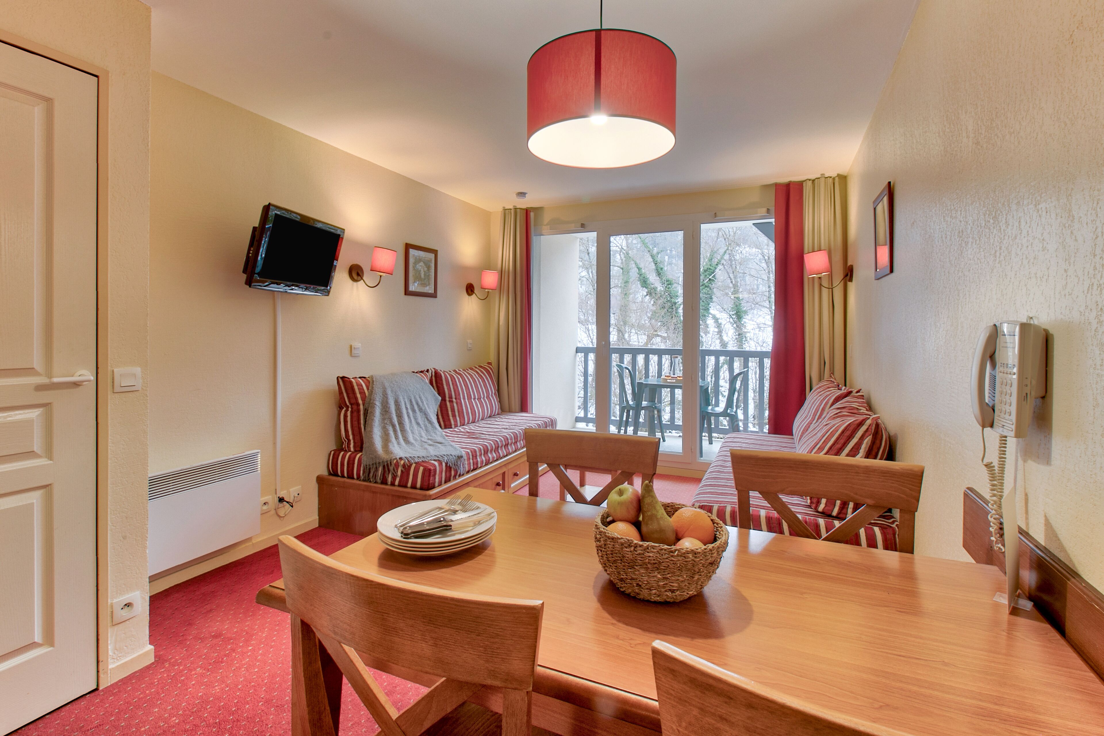 A dining area with a wooden table, chairs, and a fruit bowl. There is a television mounted on the wall, a sofa with striped cushions, and a balcony with a view.
