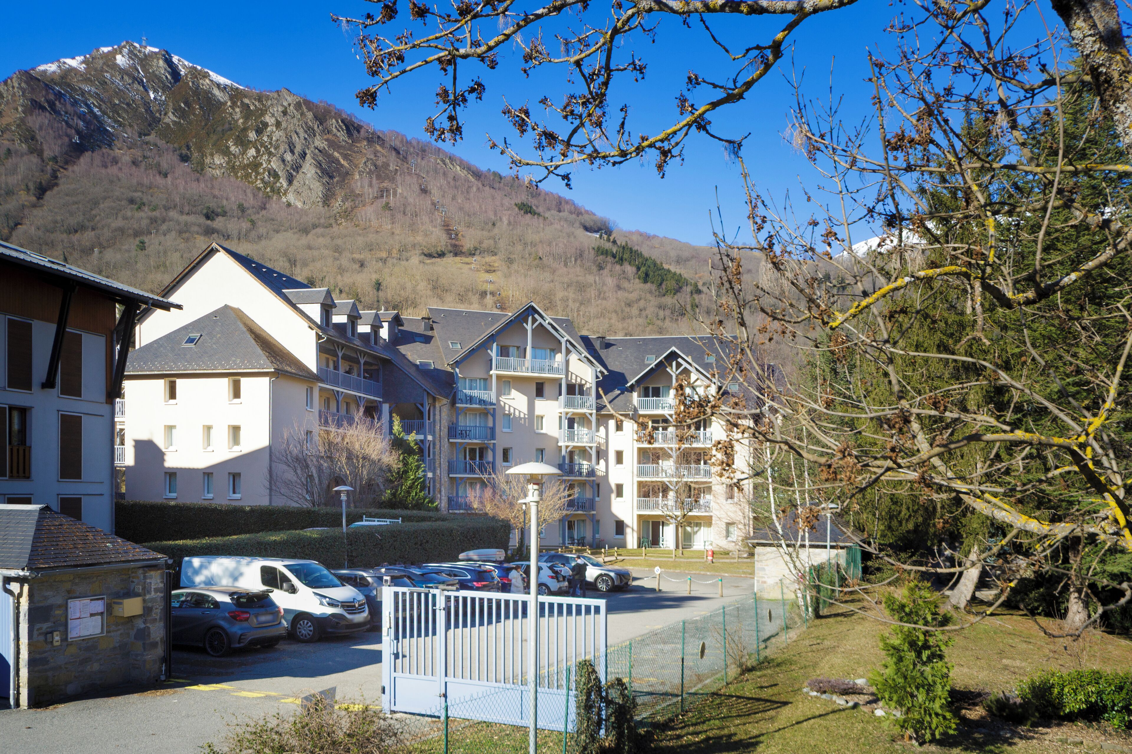 A residential building with multiple balconies, a parking area, and a mountainous backdrop.