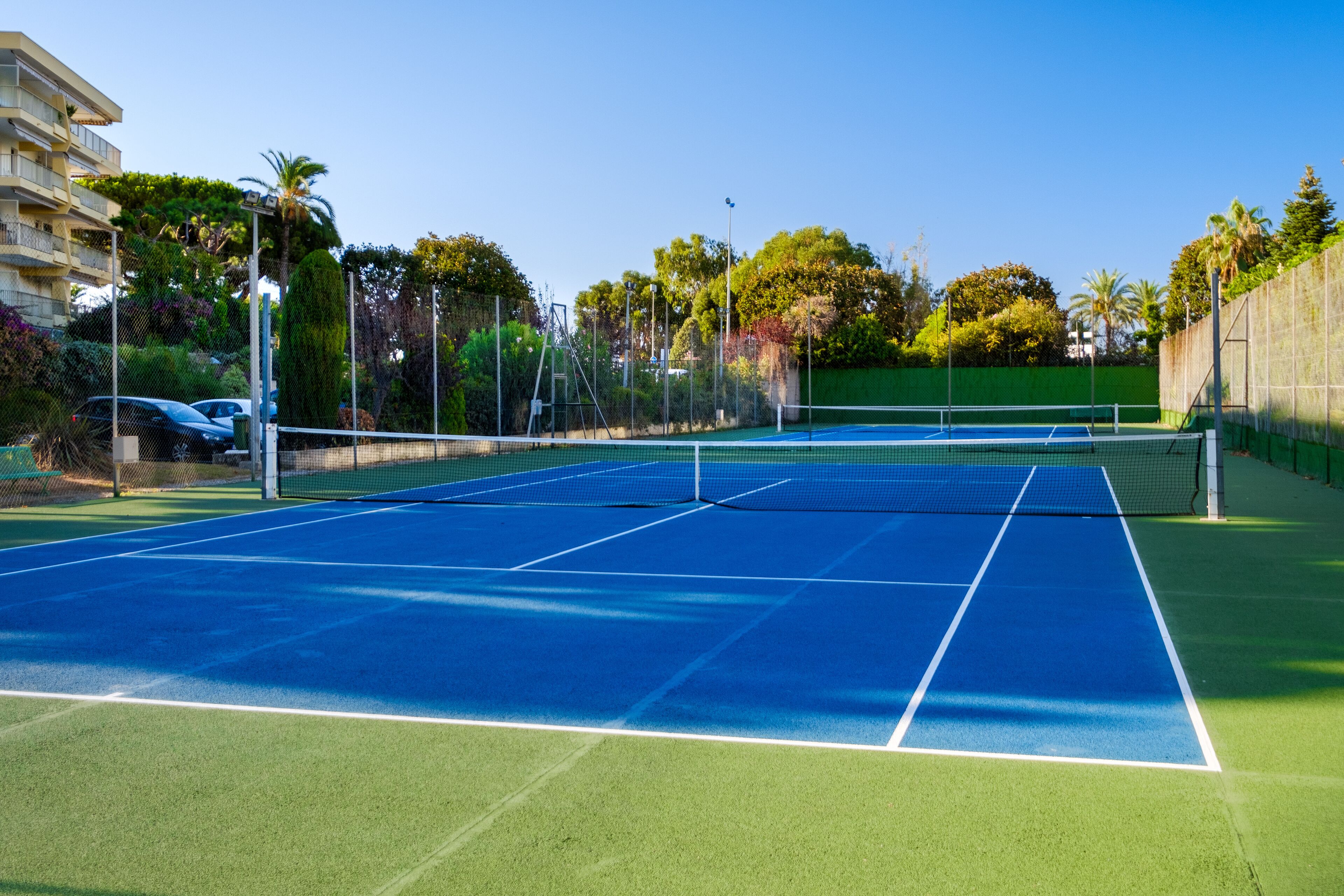 An outdoor tennis court with a net, surrounded by a fence and greenery.