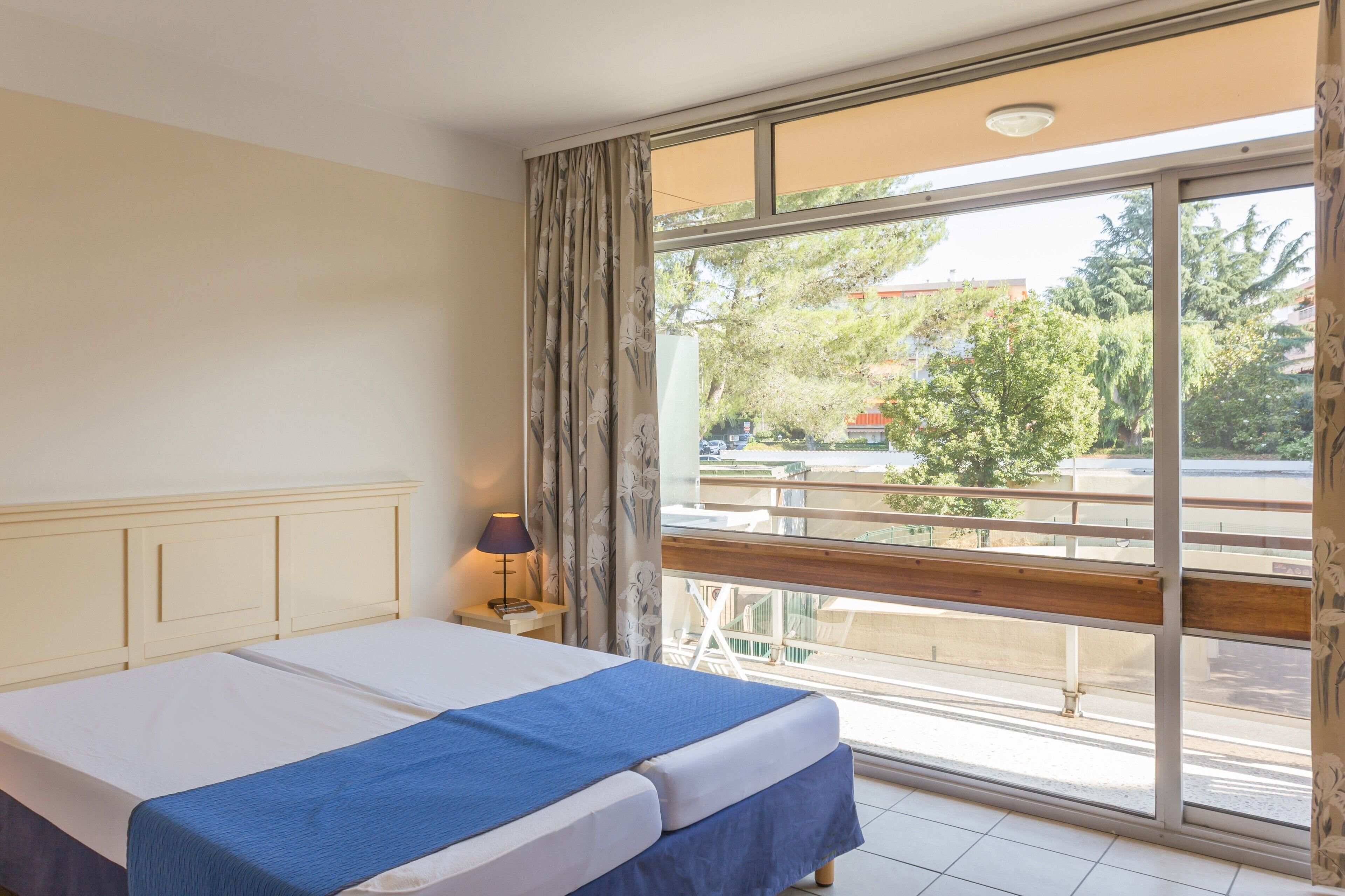 A hotel room with a bed, a nightstand, a lamp, and a large window with a view of the outside.