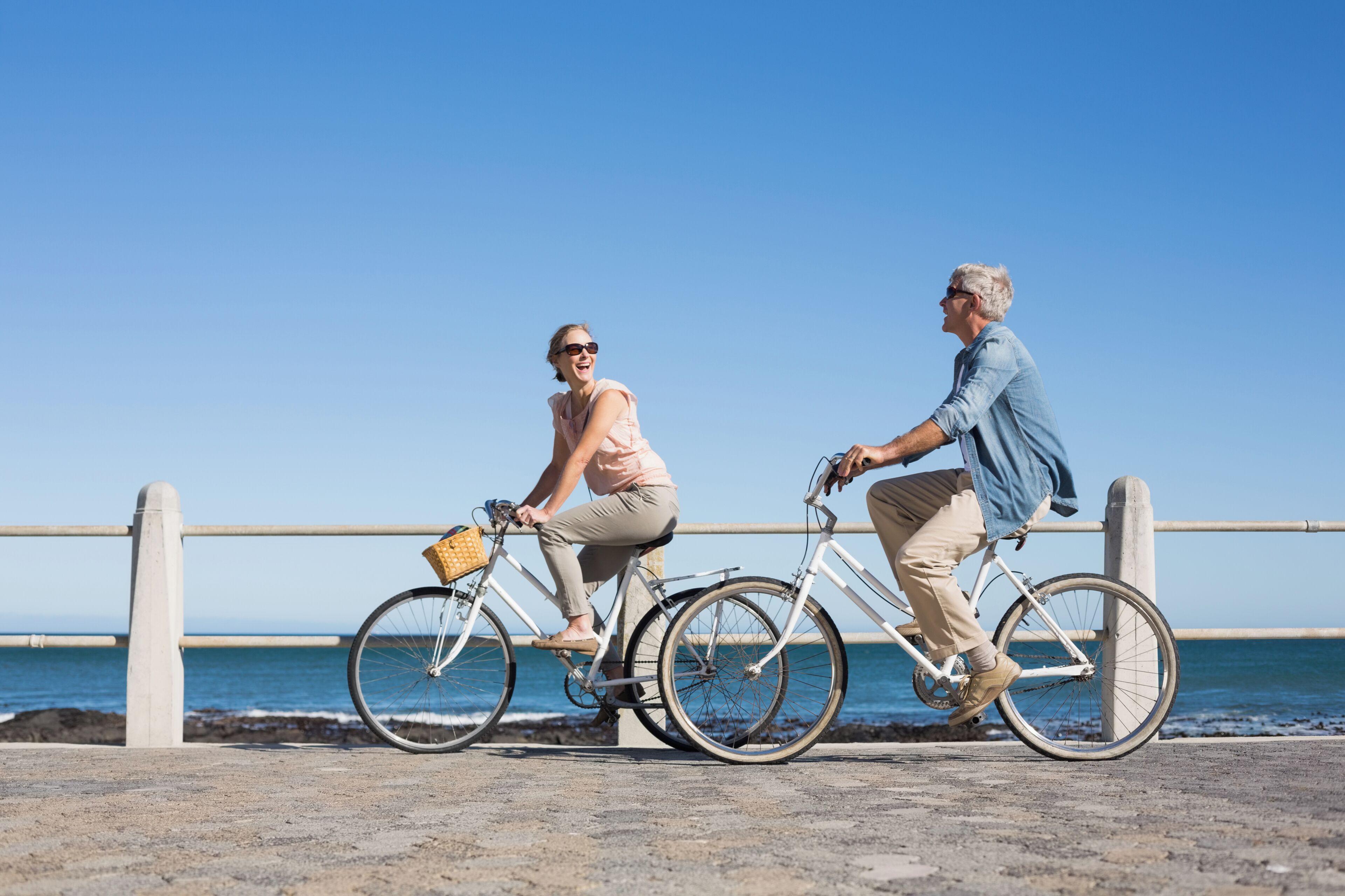 Dos personas paseando en bicicleta por un paseo marítimo con barandilla y el océano al fondo.