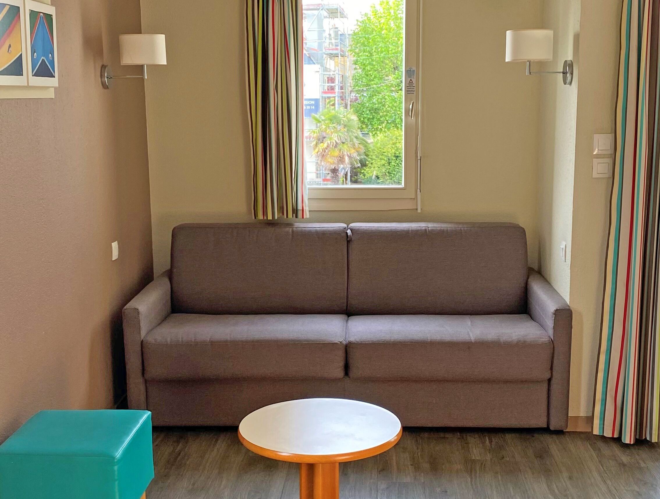 Une pièce comprenant un canapé, une petite table ronde, une fenêtre avec des rideaux et des lampes fixées au mur.