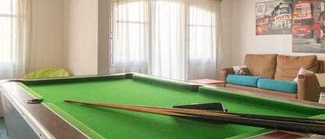 Ein Billardtisch mit grüner Filzauflage, eine Poolkeule und ein Billardständer. Im Hintergrund steht ein beiges Sofa mit einem blauen Kissen.