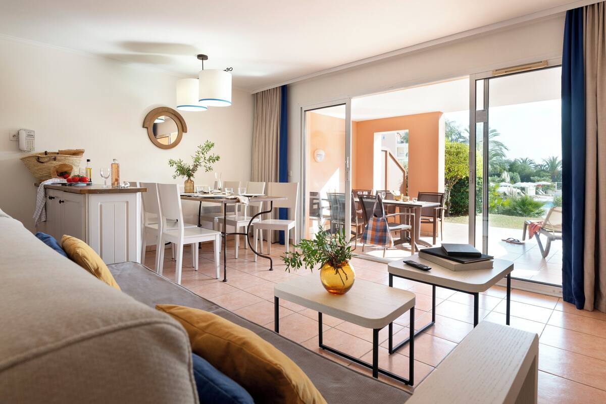 Habitación de hotel con cama, zona de comedor con sillas y mesas, pequeña cocina y vista al exterior.