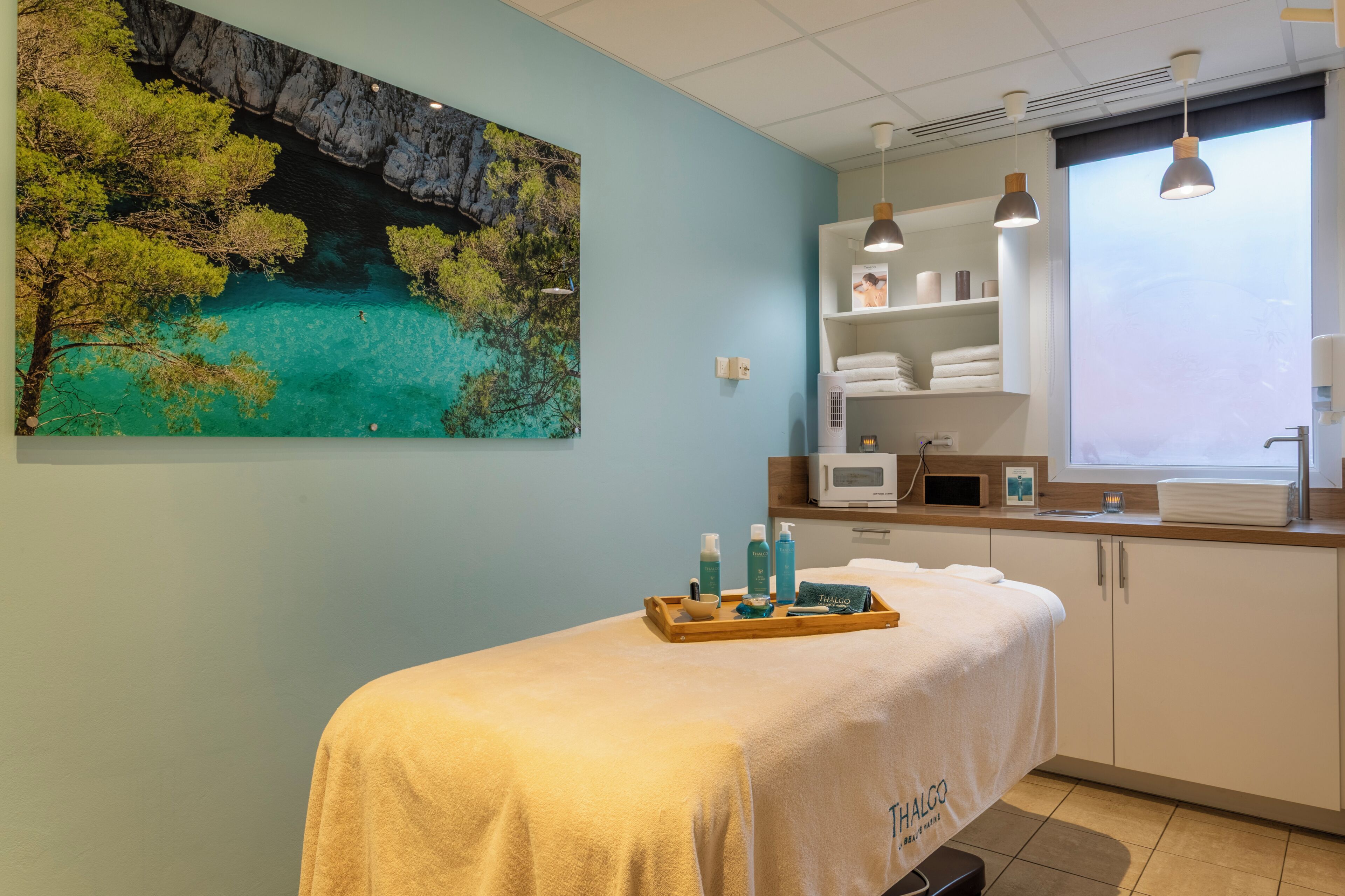 Une salle de spa équipée d’une table de massage, d’un grand tableau représentant un paysage côtier, d’une fenêtre donnant sur un beau panorama, et d’une étagère où sont rangées des serviettes et des articles de toilette.