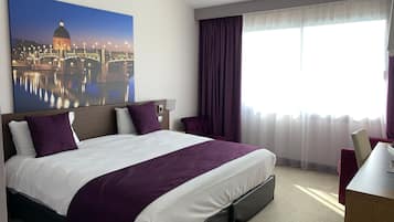 Habitación de hotel con una cama grande, un escritorio, una silla, un ventanal con cortinas y un cuadro de un paisaje urbano en la pared.