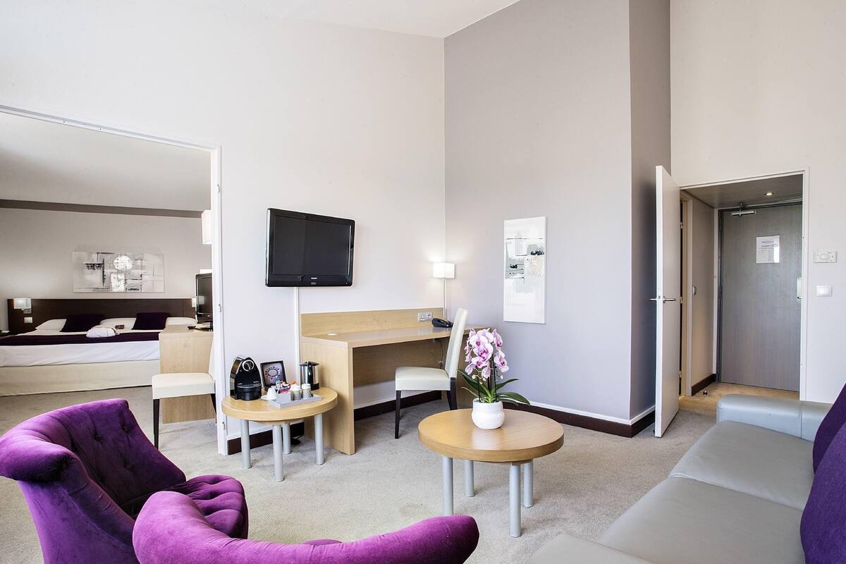 Une chambre d’hôtel avec un lit, un bureau, une télévision, une petite table ornée d’un vase de fleurs et deux fauteuils violets.