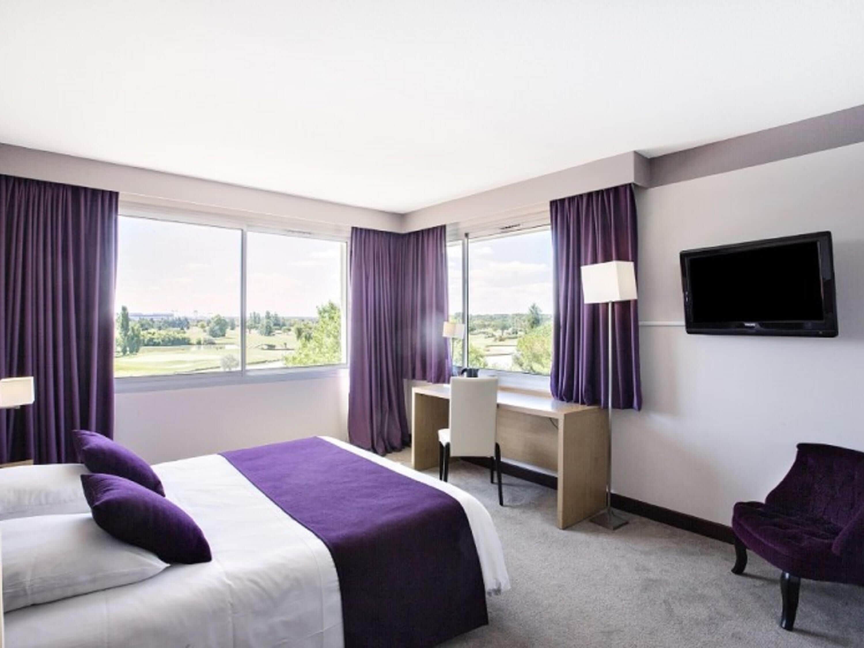 A hotel room with a bed, desk, chair, TV, and large windows with curtains.