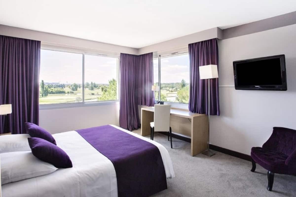 A hotel room with a bed, desk, chair, TV, and large windows with curtains.