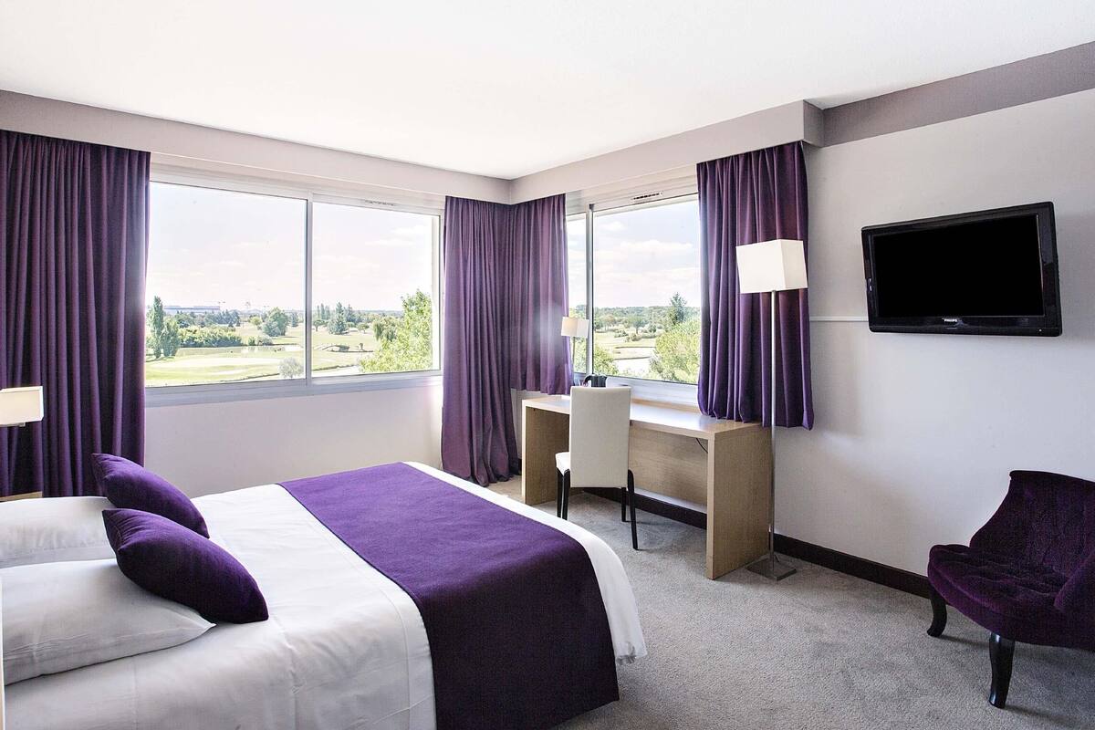 Une chambre d’hôtel équipée d’un lit, d’un bureau, d’une chaise, d’une télévision et de grandes fenêtres avec des rideaux.