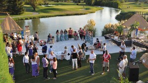 Un campo de golf con un estanque, una estructura de techo de paja y un grupo de personas reunidas alrededor de un escenario.