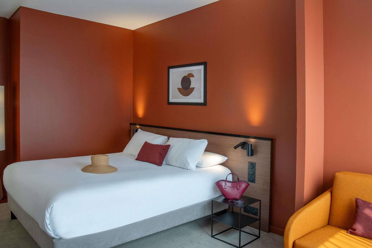 Une chambre d’hôtel moderne avec un grand lit, une table de chevet, une chaise et une œuvre d’art encadrée au mur.